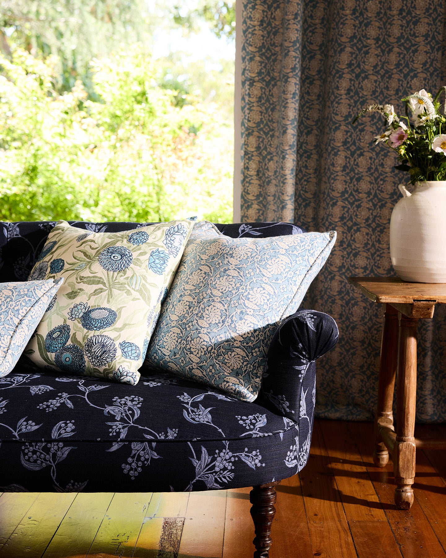 Utopia Goods: navy floral sofa with patterned throw pillows next to wooden side table with vase in sunlit room