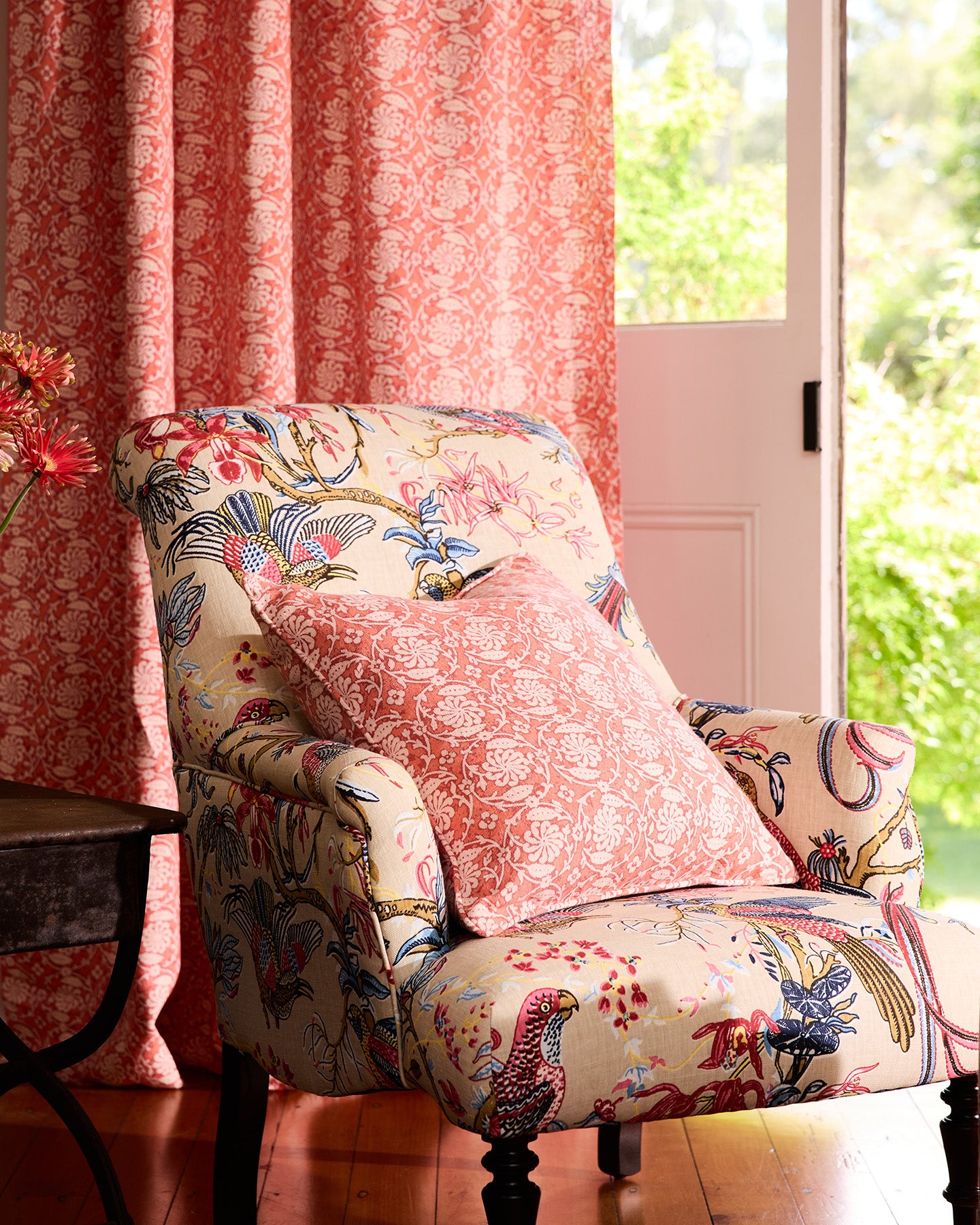 Utopia Goods: Floral armchair with coral pillow beside matching coral curtains and French doors showing garden view
