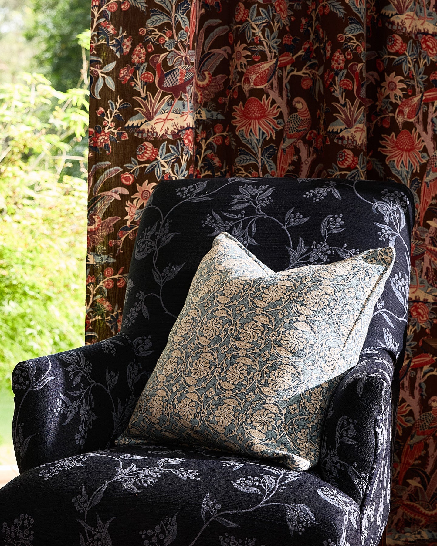 Utopia Goods: Dark floral upholstered armchair with decorative pillow next to colorful patterned curtains by window