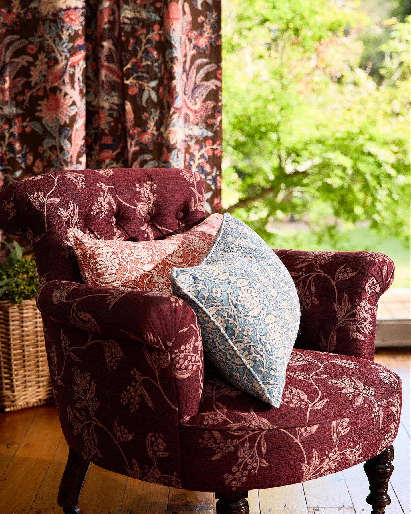 Utopia Goods: burgundy floral armchair with colorful throw pillows beside a window overlooking green garden foliage