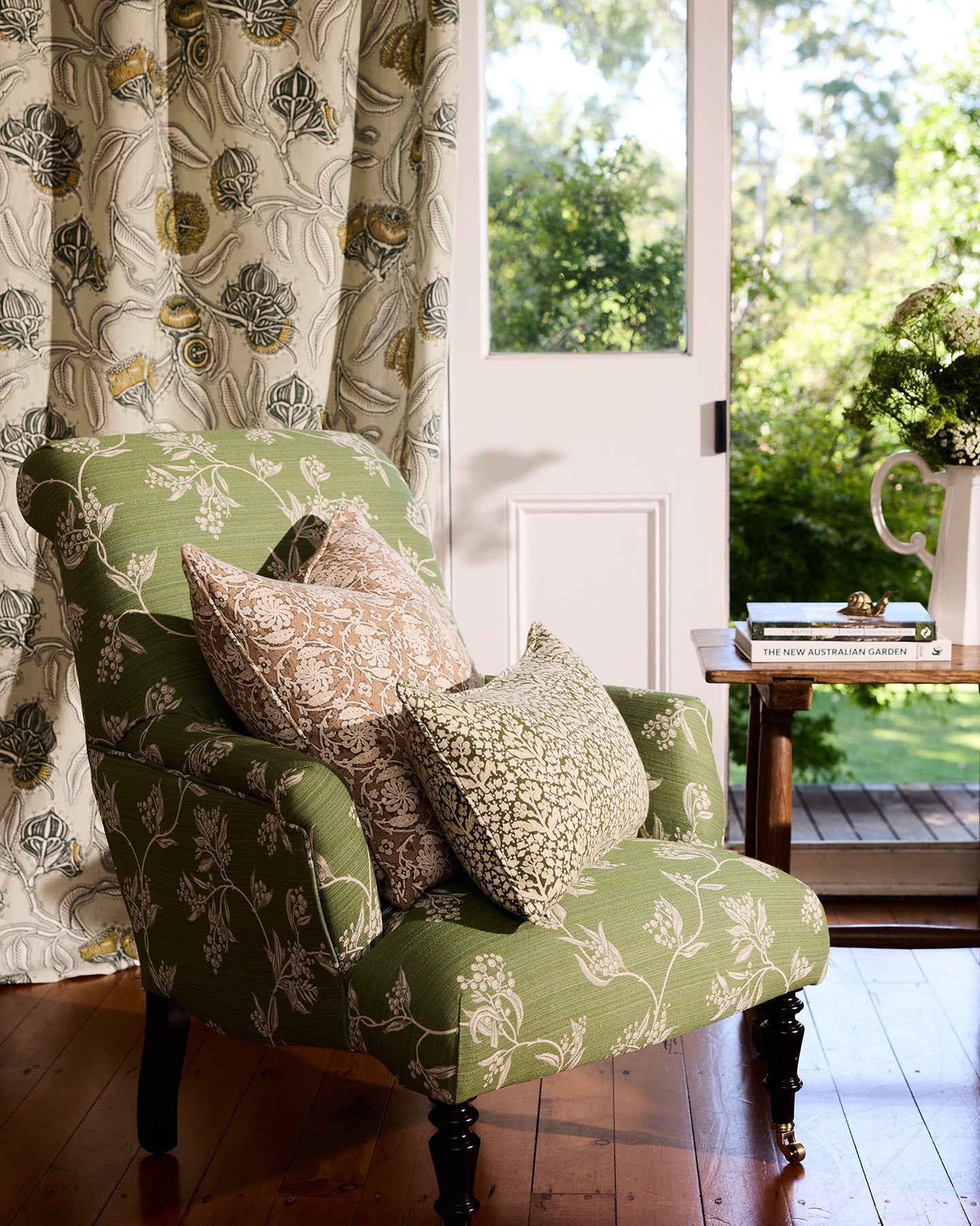 Utopia Goods: Green floral armchair with decorative pillows next to wooden side table, white door open to garden view