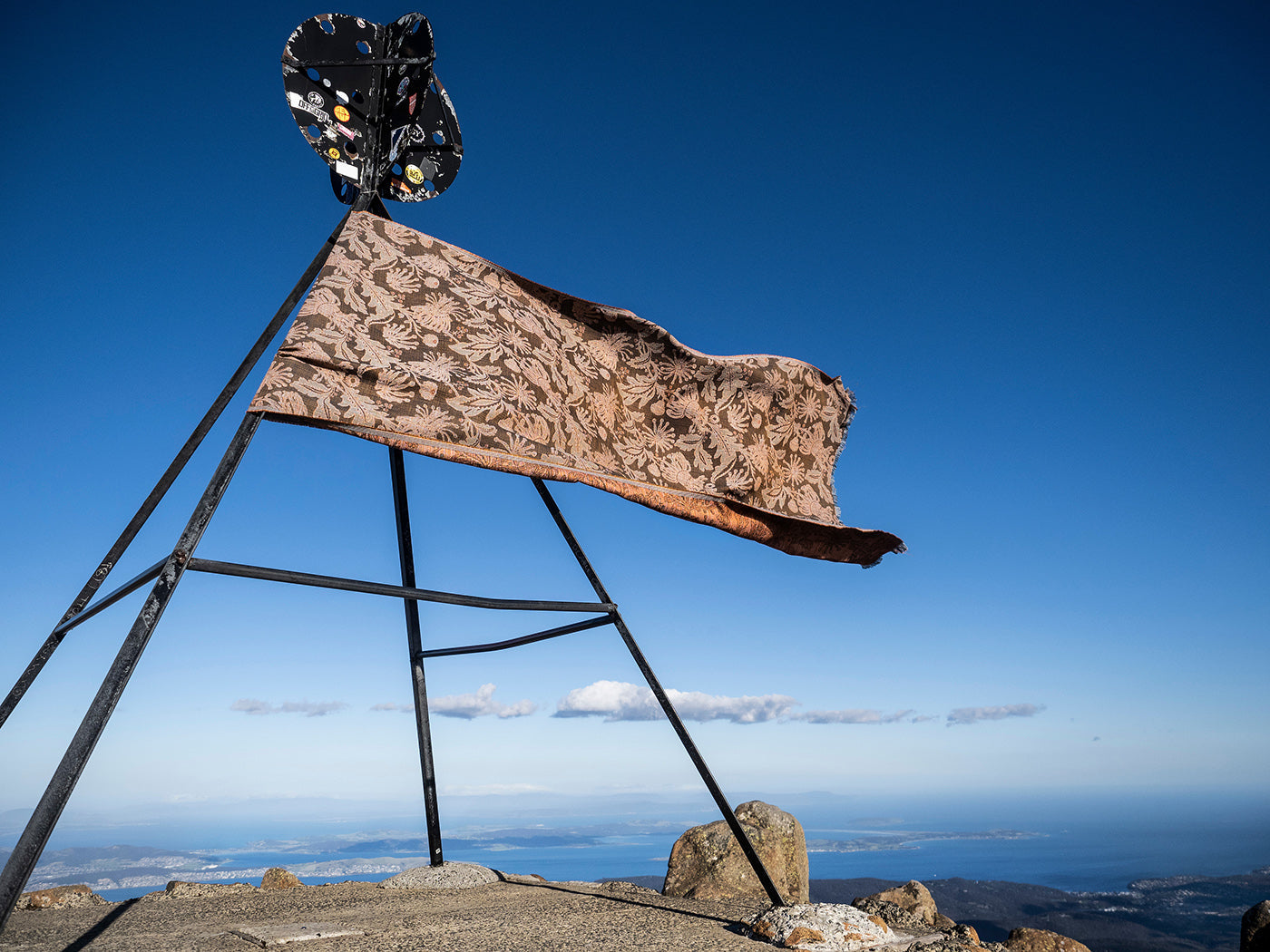 Utopia Goods Firewheel Rust Performance Fabric: rust-colored fabric with botanical print draped on metal frame against blue sky
