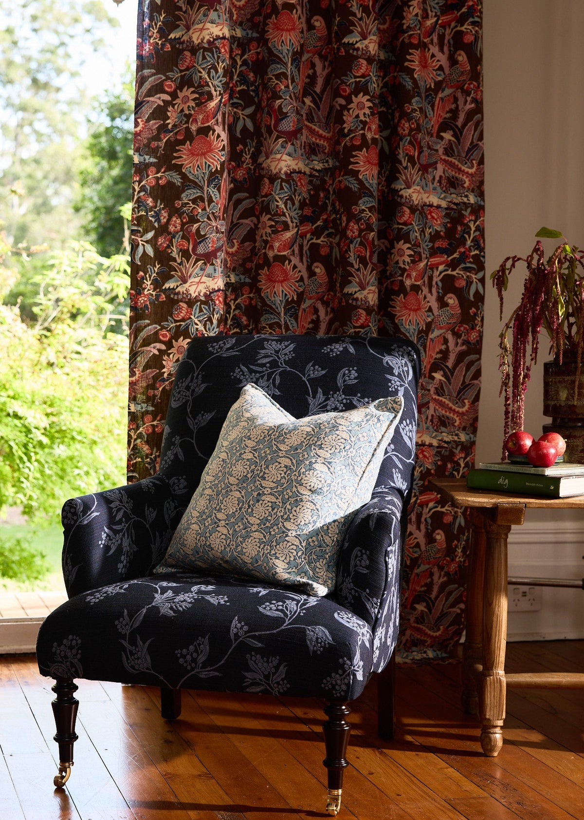 A Wattle blue floral performance fabric armchair with a light blue and white patterned Pinwheel Ocean throw pillow, set in a sunlit room with a window, Arcadia Mocha curtains, and a wooden floor.